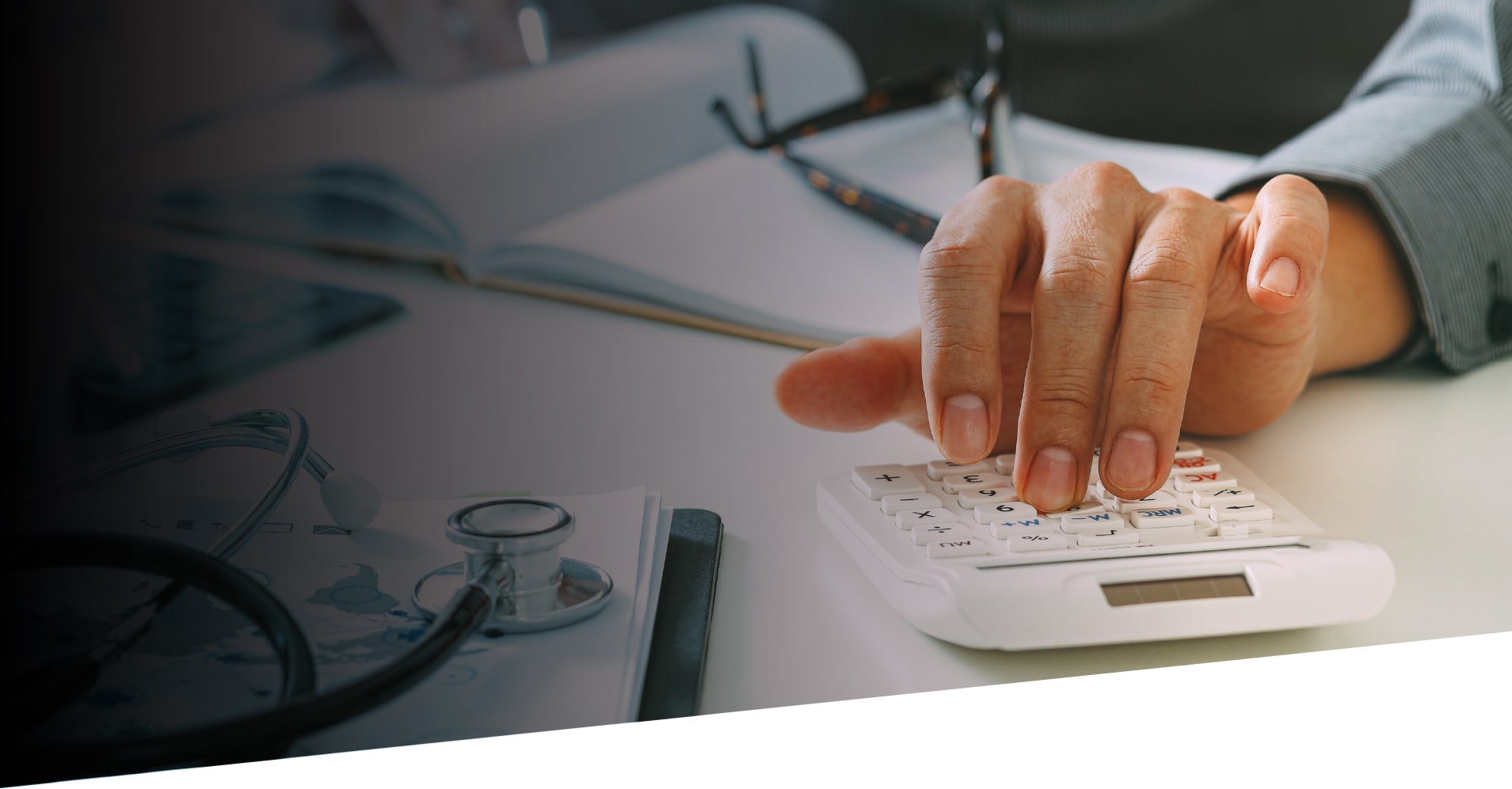 A hand using a calculator with various doctors tools on the surrounding table